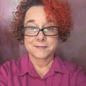 portrait of a woman with curly hair, dyed orange in the front and wearing rectangular black rimmed glasses and a pink blouse