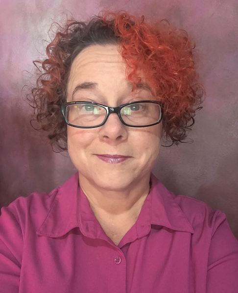 portrait of a woman with curly hair, dyed orange in the front and wearing rectangular black rimmed glasses and a pink blouse