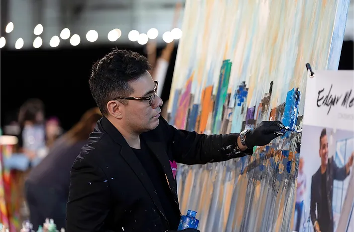 man dark, short curly hair wearing glasses painting a large canvas in a festive atmosphere
