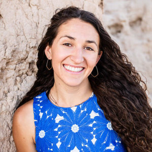 Smiling young woman with long wavy brown hair in a patterned blue dress next to a textured wall