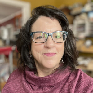 middle aged woman with brown hair and glasses, smiling at the camera while wearing a reddish pink cowl neck sweater