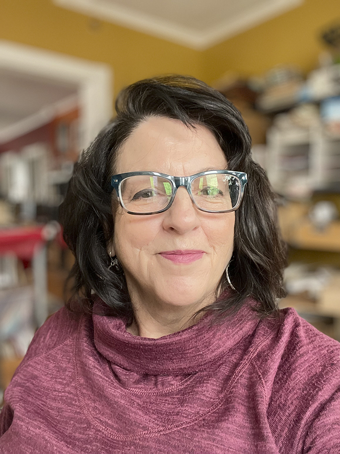 middle aged woman with brown hair and glasses, smiling at the camera while wearing a reddish pink cowl neck sweater